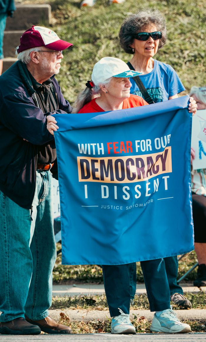 Home Three seniors hold a protest banner stating 'With Fear for Our Democracy I Dissent' outdoors.
